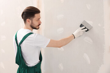 Worker in uniform plastering wall with putty knife indoors