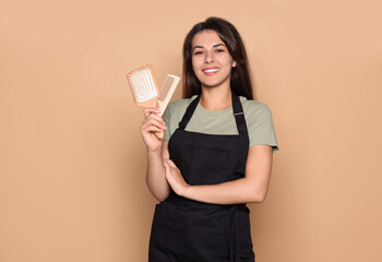 Happy professional hairdresser in apron with hairbrush and comb against pale orange background