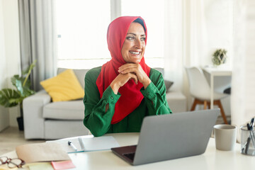 Positive islamic woman in hijab sitting at desk with laptop at home and thinking about something and smiling