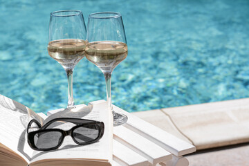Glasses of tasty wine, sunglasses and open book on swimming pool edge. Space for text