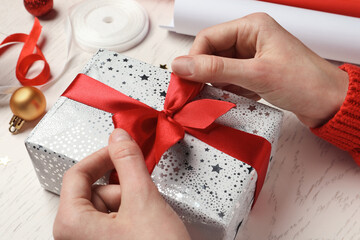 Christmas present. Woman tying ribbon bow on gift box at white wooden table, closeup