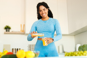 Positive black fit woman enjoying tasty lunch and pouring orange juice from jug to glass, standing...