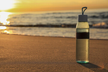 Glass bottle with water on wet sand near sea at sunset. Space for text