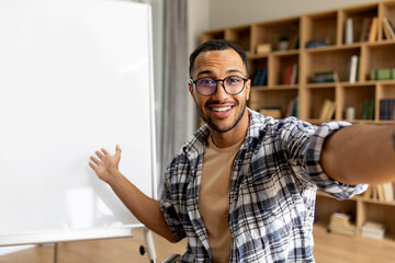 Cheerful arab tutor man smiling to camera and pointing at whiteboard, having online lesson in classroom