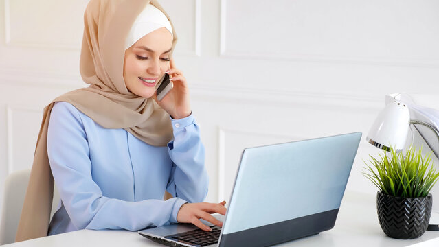 Young Beautiful Muslim Woman In Beige Hijab And Blue Dress Works In Office, Talking Mobile Phone And Typing On Laptop At Her Workplace.