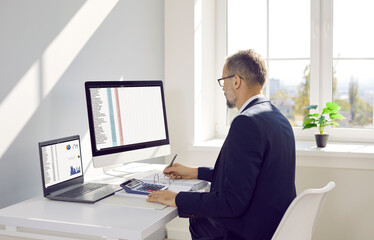 Business accountant working in office. Serious busy mature man in suit and glasses sitting in front of desktop and laptop computers, working with data, analyzing information, taking notes on paper