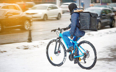 Food delivery worker on bike delivering food at snowy winter day. Delivery person with backpack cycling through blizzard. Restaurants delivery service ride through snow road. Takeaway courier.