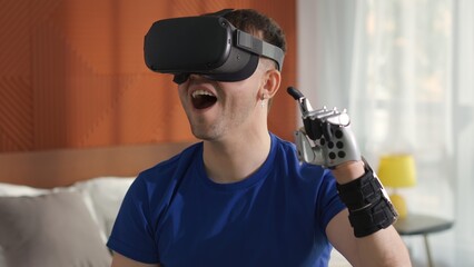Young man contemporary bionic arm use virtual reality headset playing video game sitting on bed 