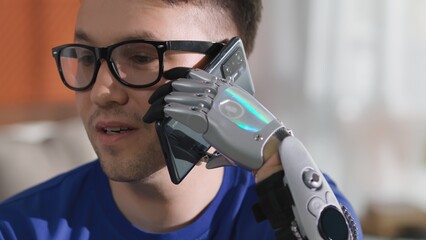 Young disabled man with bionic arm is holding phone and talking sitting at home 