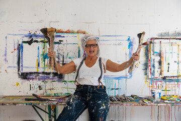older, gray-haired, mature, loudly laughing, happy artist with glasses stretches the large brushes upwards in a leap of joy