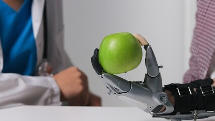Close up of man with robotic arm prosthesis holds green apple at doctor's office