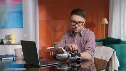 Portrait of young handicapped man sit down at table in living room and remove arm prosthesis