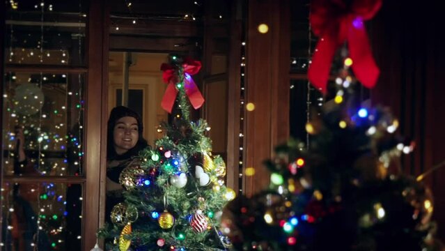 A young girl stands outside the window and looks at a beautiful Christmas tree through the glass. Christmas mood. Beautiful and rich decorated Christmas fir tree in the european home. Close up a