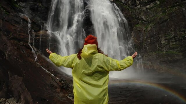 Young girl traveler hiker in a yellow raincoat walks to a waterfall in the highlands, raises his hands up, enjoys nature and life. Traveling in the mountains, adventure in trip. Lifestyle concept