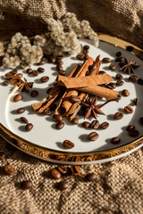 An elegant composition of candles, plates with a golden border, on which spices - anise, cinnamon and coffee beans lie. Against the background of burlap