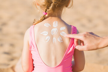 Young adult mother hand applying sunscreen lotion on little girl back. Sun shape created from white cream. Skin protection. Safety sunbathing in hot sunny day at beach. Rear view. Closeup.