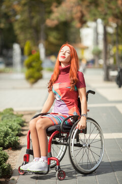 Beautiful red-haired girl on a wheelchair in a city park. Walks in the open air. Socialization of the disabled. accessible environment.