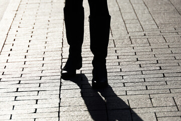 Female legs in boots on a street, black silhouette and shadow of woman walking on paved sidewalk