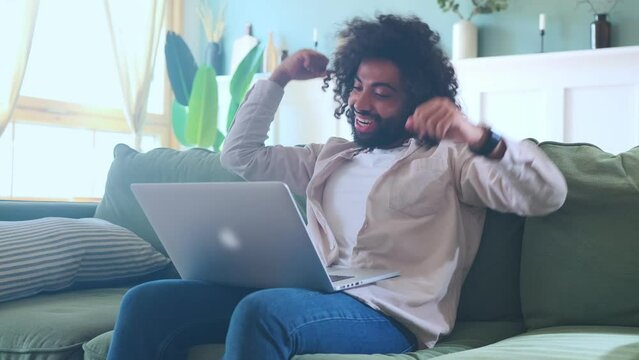 Young overjoyed Arabian man with laptop on knees makes victorious wave of hands after learning about victory of sports team on which bet was placed at bookmakers sits on sofa in living room