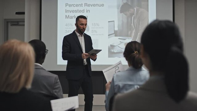 Moden young adult businessman with beard on face wearing microphone holding digital tablet starting lecture about investment