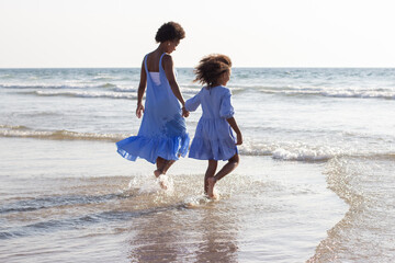 Back view of tender mother and daughter on vacation. African American family in beautiful dresses walking in waves, having good time, holding hands. Family, travelling, parenthood concept