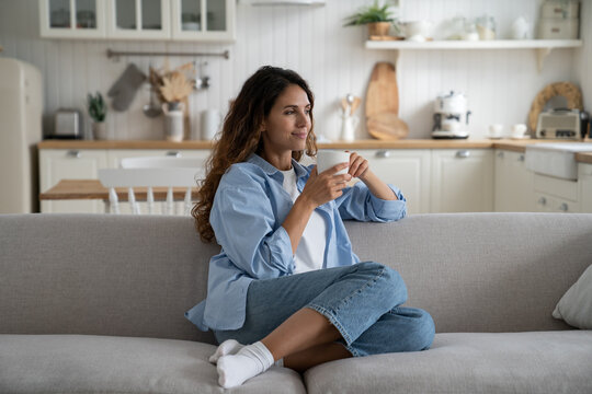 Cheerful Spanish woman with mug filled with tea or coffee looks into distance and smiles sincerely remembering college studies or friends from university. Casual Caucasian girl sits alone on gray sofa