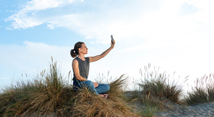 Caucasian woman sitting on a sand dune taking a selfie on a mobile phone.