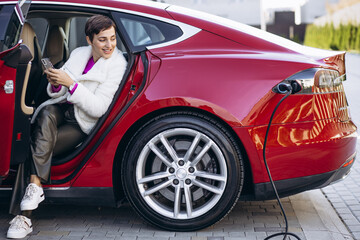 Naklejka na ściany i meble Woman charging electric car and talking on the phone