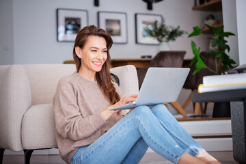 Fototapeta na wymiar Smiling caucasian woman using laptop at home while sitting on the floor