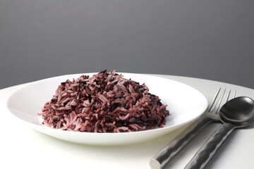 cooked brown rice berry rice (non-glutinous purple rice) in the white bowl with stainless steel spoon and fork are on the white steel table of the organic food restaurant with grey wall background
