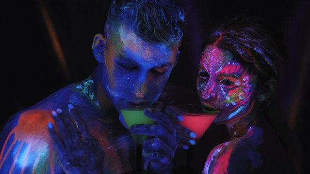 A couple with UV body art dancing and clinks glasses of colorful glowing cocktails in the dark under the light of fluorescent lamps. Art design of couple disco dancers posing in UV.