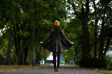 Female standing in olive coat, yellow hat and red shoes on an alley with green trees during autumn