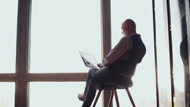 man working on a laptop sitting on a chair in front of the window.