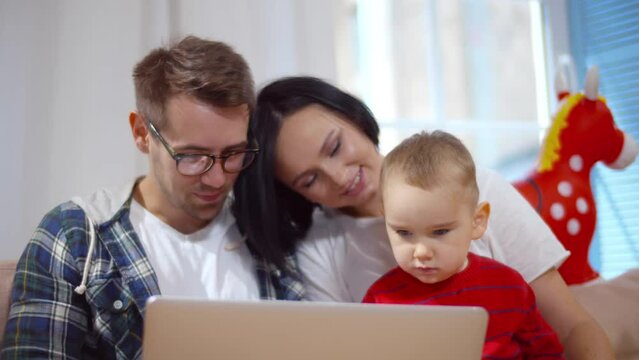 Portrait of caucasian family use laptop sitting on couch with baby at home. Realtime