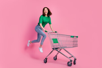 Full body photo of carefree overjoyed lady jumping push supermarket trolley isolated on pink color...