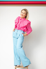 Joyful adult woman in bright pink blouse and blue trousers standing on chair, isolated on white. Full length photo of confident successful business lady. Happy people, fashion clothes