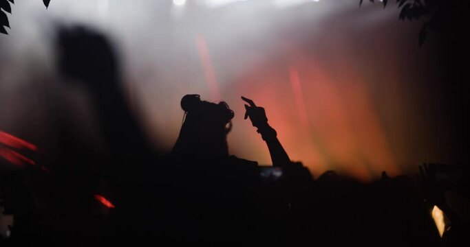 Silhouette people in front of a stage with flashing lights in crowd of people with hands up holding phones and dancing.