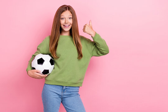 Photo of confident shiny school girl dressed green sweatshirt holding football ball thumb up empty space isolated pink color background - Powered by Adobe