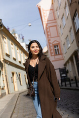 Cheerful woman in brown coat looking away on street with sunlight in Prague.