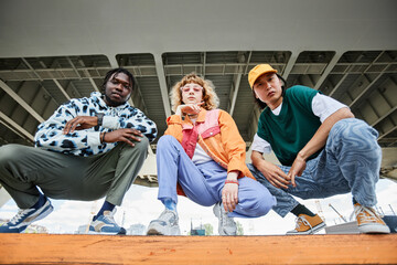 Group of three young people wearing street style clothes outdoors while sitting on stairs in urban...