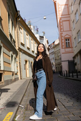 Fototapeta premium Full length of stylish woman in coat walking on road between buildings on street in Prague.