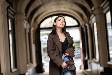 brunette woman in coat holding handbag and looking away near arch building in prague.