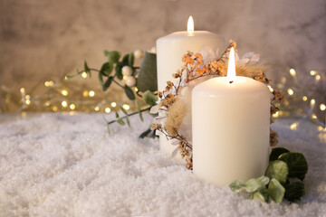 Two white candles decorated in Christmas decor on the snow against the background of lights