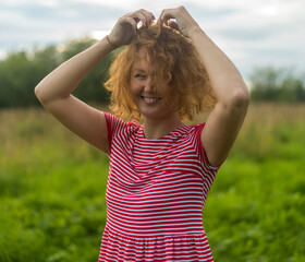 a girl in a red summer dress with loose red hair on the background of a green meadow and a sky with clouds