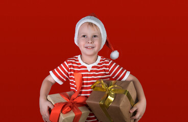 Funny smiling joyful baby boy in a red Santa hat holding a Christmas gift in his hand on a red background. Layout with copy space