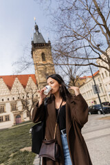 Stylish young woman drinking takeaway coffee on urban street in Prague.