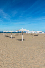 sunny sandy beach with numerous white wooden umbrellas against a blue cloudy sky