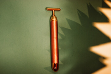 Vibrating golden facial massager, glass jar with oil dropper for massage on emerald background....