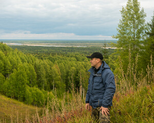 Naklejka na ściany i meble a man in a blue jacket and a black cap on a wooded hill against the background of a forest and a river.