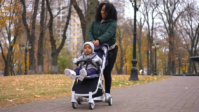 Portrait of smiling mother rolling baby stroller with toddler son smiling walking along park alley. Wide shot happy African American woman enjoying weekend leisure with cute boy on autumn day outdoors